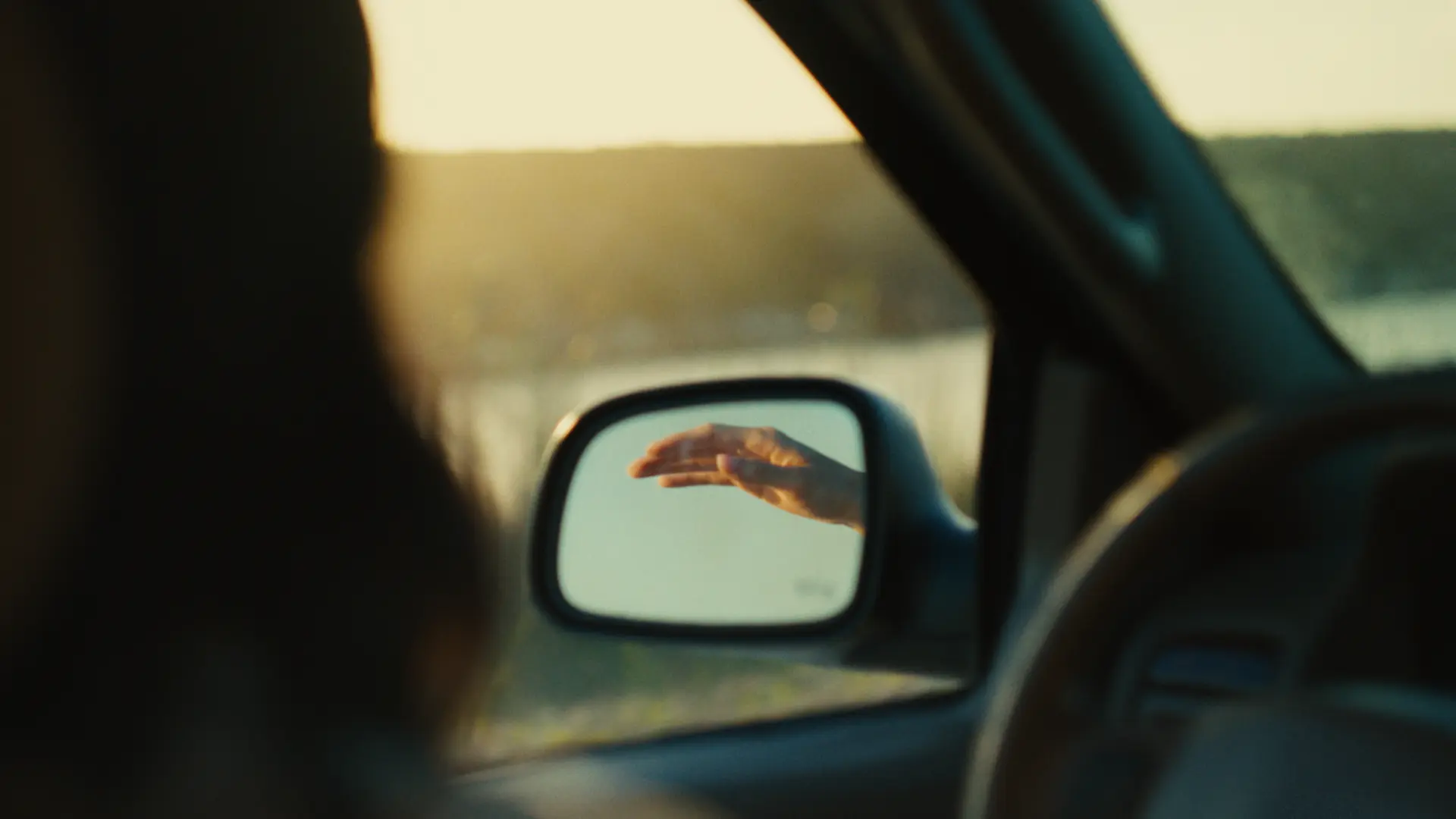 A hand reflected in a car side mirror at golden hour on an open road